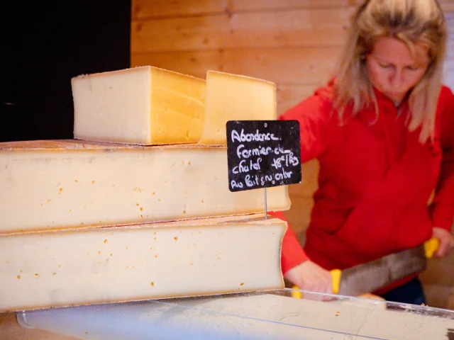 Premier plan : un abondance fermier. Second plan : une dame habillé en rouge qui coupe du fromage.