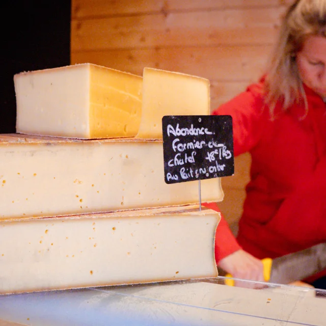 Premier plan : un abondance fermier. Second plan : une dame habillé en rouge qui coupe du fromage.