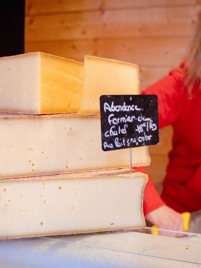 Premier plan : un abondance fermier. Second plan : une dame habillé en rouge qui coupe du fromage.