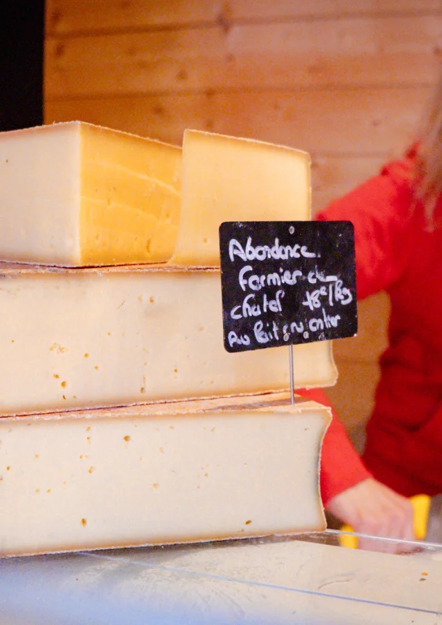 Premier plan : un abondance fermier. Second plan : une dame habillé en rouge qui coupe du fromage.