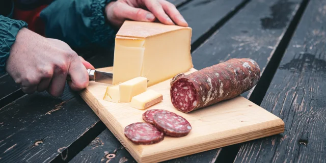 Une planche en bois sert de support à un saucisson pré-découpé tandis que quelqu'un coupe des tranches d'Abondance avec un Opinel.