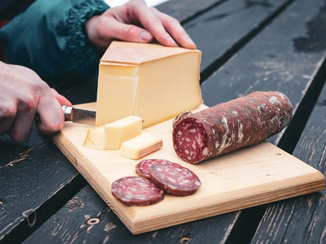 Une planche en bois sert de support à un saucisson pré-découpé tandis que quelqu'un coupe des tranches d'Abondance avec un Opinel.