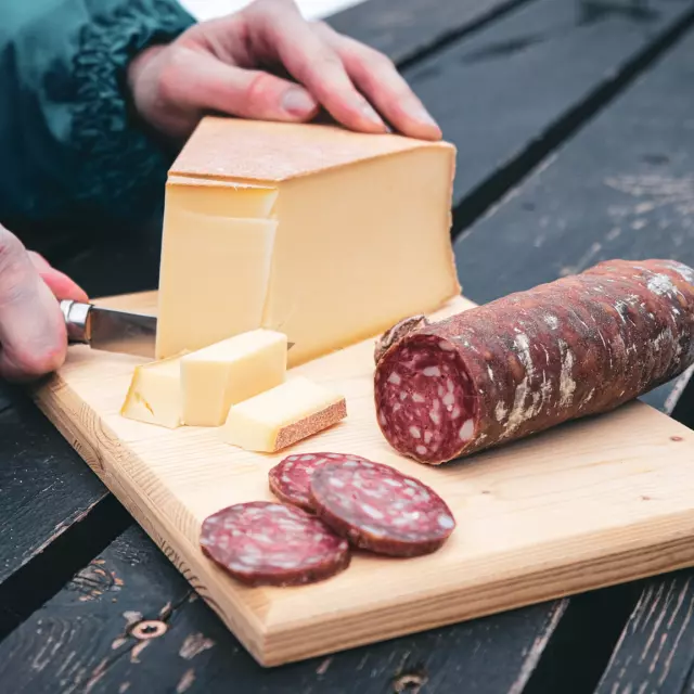 Une planche en bois sert de support à un saucisson pré-découpé tandis que quelqu'un coupe des tranches d'Abondance avec un Opinel.