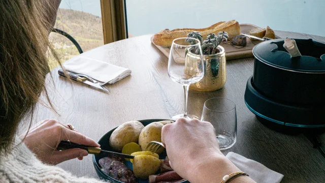 Fondue sur une table en bois, une jeune femme coupe ses pommes de terre avec vue sur le Leman