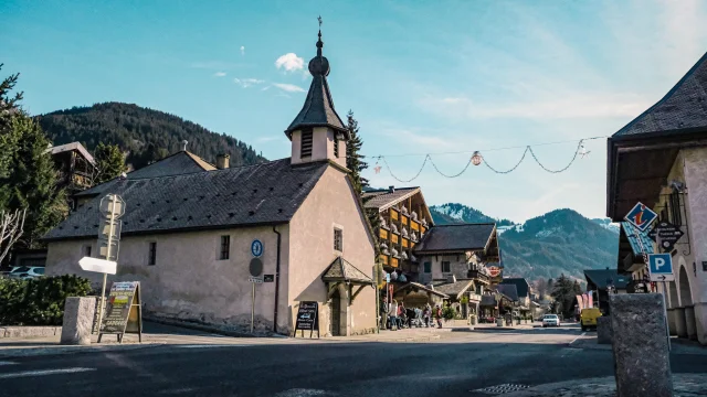 Village La Chapelle D Abondance. Une petite Chapelle se trouve au premier plan. Une route traverse le village, une voiture arrive en fond et il y a un groupe sur la gauche.