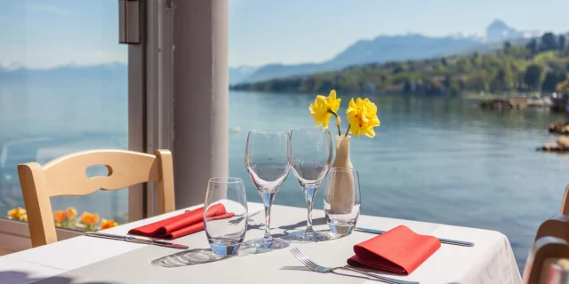 Table nappe blanche, vue sur le lac, deux verres de vin vides et deux fleurs jaunes dans un vase