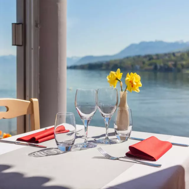 Table nappe blanche, vue sur le lac, deux verres de vin vides et deux fleurs jaunes dans un vase