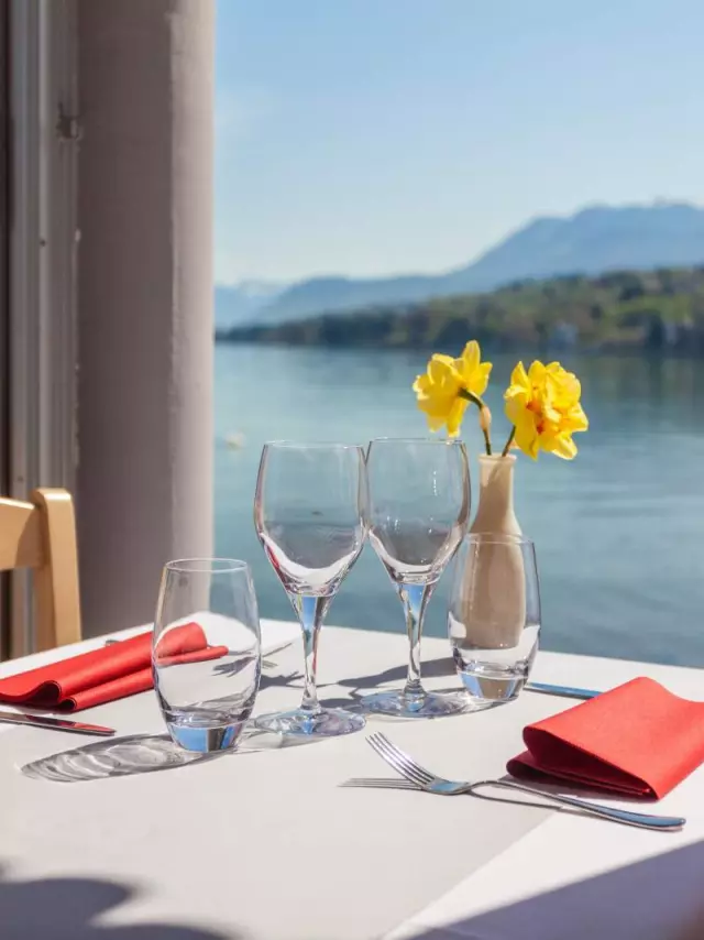 Table nappe blanche, vue sur le lac, deux verres de vin vides et deux fleurs jaunes dans un vase