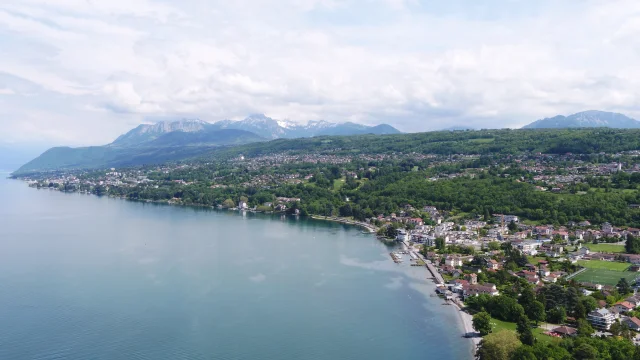 Lac et côte en premier plan. On voit les bâtiments et les arbres grâce à une vue drône avec en fond les montagnes