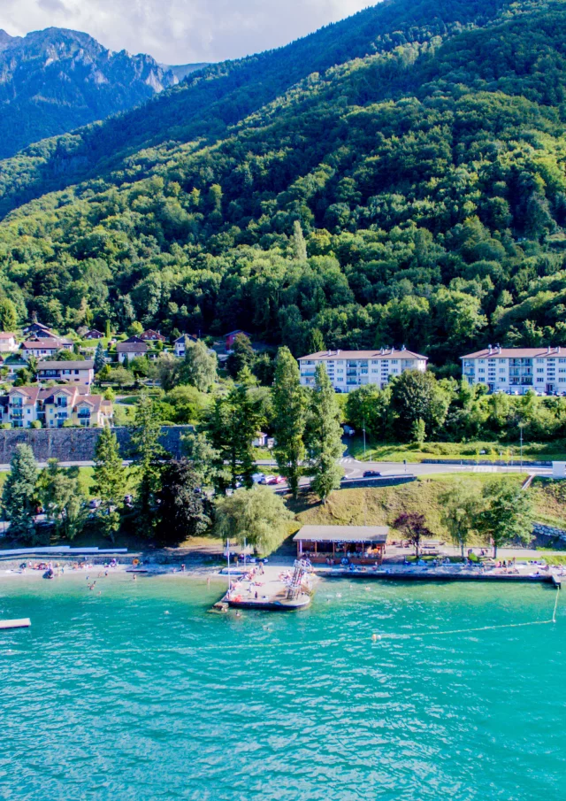 Vue drône depuis le lac. L'eau est bleu et on voit la montagne en fond avec le port.